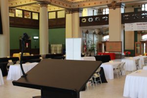 The rotunda within 181 Higgins, populated with tables and chairs for an event. Photo has a lectern focused and in the foreground is seating for an event