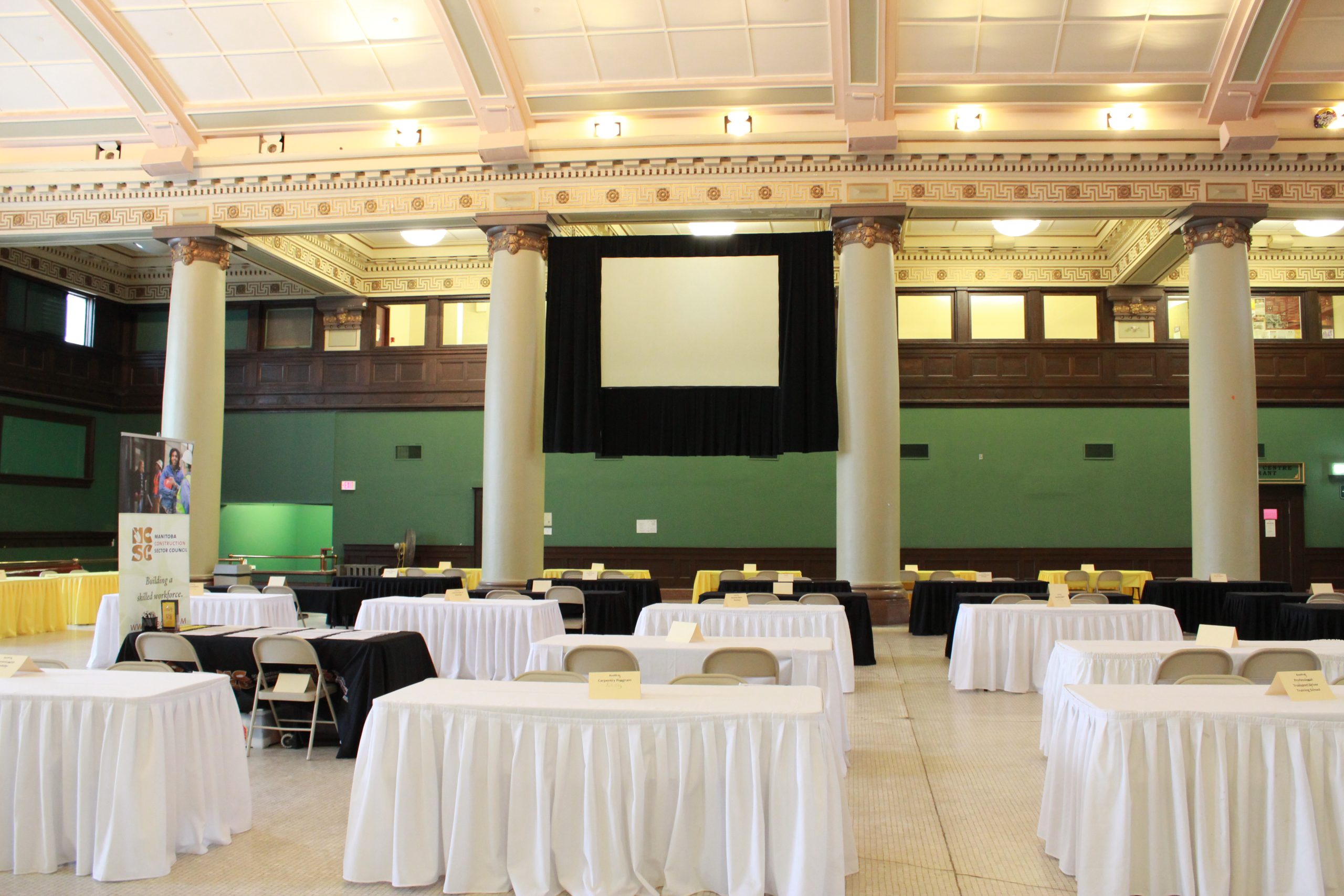 The rotunda within 181 Higgins, populated with tables and chairs for an event. Photo is facing towards a projector screen on the eastern wall